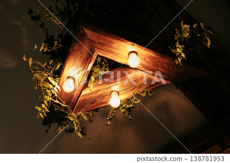 Decorative wooden ceiling light fixture with warm bulbs and green plants. Concept of interior design, ambiance lighting and cozy atmosphere Decorative wooden ceiling light fixture with warm bulbs and green plants. Concept of interior design, ambiance lighting and cozy atmosphere 138781953