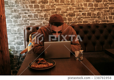 Man working on laptop in cafe with food on table in warm cozy interior. Concept of remote work, digital lifestyle and modern urban routine Man working on laptop in cafe with food on table in warm cozy interior. Concept of remote work, digital lifestyle and modern urban routine 138781958