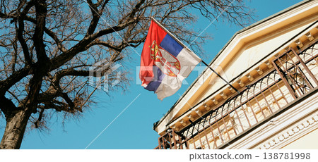 Serbian flag waving near historic building and tree branches against blue sky. Concept of patriotism, identity and cultural heritage in urban environment Serbian flag waving near historic building and tree branches against blue sky. Concept of patriotism, identity and cultural heritage in urban environment 138781998