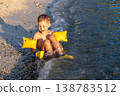 Happy child sits sandy beach shoreline small waves splash wearing bright inflatable armbands under warm golden sunlight. Playful summer vacation scene joy freedom leisure carefree childhood by sea 138783512