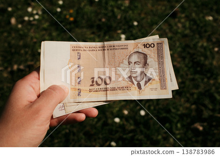 Hand holding Bosnia and Herzegovina convertible mark banknote outdoors against green grass background. Financial concept of cash payment, currency value, and personal finance 138783986