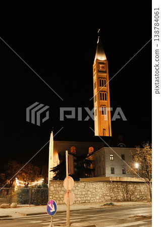 Night view of a Catholic church tower illuminated above surrounding buildings in Mostar, with warm lights and a dark sky emphasizing historic urban architecture Night view of a Catholic church tower illuminated above surrounding buildings in Mostar, with warm lights and a dark sky emphasizing historic urban architecture 138784061