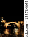Night view of the Old Bridge in Mostar with illuminated stone arch and medieval towers reflecting in the Neretva River under a dark sky 138784065