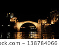 Night view of the Old Bridge in Mostar with illuminated stone arch and medieval towers reflecting in the Neretva River under a dark sky 138784066