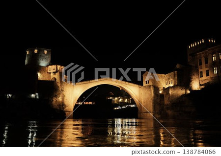 Night view of the Old Bridge in Mostar with illuminated stone arch and medieval towers reflecting in the Neretva River under a dark sky 138784066