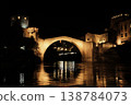 Night view of the Old Bridge in Mostar with illuminated stone arch and medieval towers reflecting in the Neretva River under a dark sky 138784073