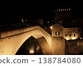 The iconic stone bridge of Mostar glows under night lighting above the river. Historic architecture and dramatic contrast highlight the landmark against the dark sky 138784080