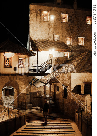 Night view of historic stone buildings in Mostar old town, illuminated with warm lights, showing traditional architecture, wooden roofs, stairway and a single person standing on old bridge. Night view of historic stone buildings in Mostar old town, illuminated with warm lights, showing traditional architecture, wooden roofs, stairway and a single person standing on old bridge. 138784081