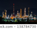 Workers operate machinery and equipment at an oil refinery during the night near hills and urban areas. Lights shine brightly 138784730