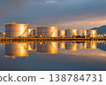 Several oil storage tanks are lined up beside the water, showing their reflection as the sun sets in the background 138784731