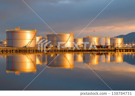 Several oil storage tanks are lined up beside the water, showing their reflection as the sun sets in the background Several oil storage tanks are lined up beside the water, showing their reflection as the sun sets in the background 138784731