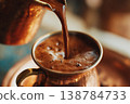 Hot coffee is poured from a metallic pitcher into a brass cup in a warm and inviting space during the morning 138784733