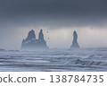 Waves crash on a beach as tall rocks rise from the sea under a cloudy sky with birds flying above 138784735