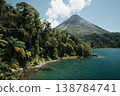 A tall volcano rises next to a clear blue lake surrounded by green trees under a bright sky with some clouds 138784741