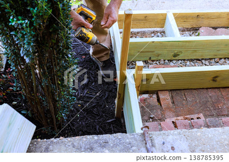 Man uses drill to secure wood steps frame near bush in garden area Man uses drill to secure wood steps frame near bush in garden area 138785395
