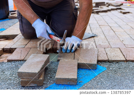 Worker lays bricks on path while wearing gloves and using hammer during construction project Worker lays bricks on path while wearing gloves and using hammer during construction project 138785397