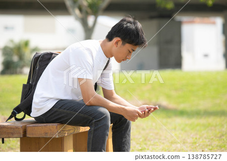 A male student is using his smartphone on a park bench. 138785727