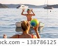 Happy child plays calm sea water inflatable ring mother throws colorful ball warm summer vacation. Bright warm sunlight waves yachts create joyful family atmosphere filled laughter carefree moments 138786125