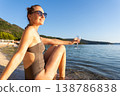 Smiling woman in sunglasses relaxes near calm water holding glass during warm sunset. Natural beauty, summer vacation mood, peaceful landscape freedom leisure serene travel atmosphere by quiet shore 138786838
