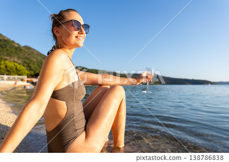 Smiling woman in sunglasses relaxes near calm water holding glass during warm sunset. Natural beauty, summer vacation mood, peaceful landscape freedom leisure serene travel atmosphere by quiet shore Smiling woman in sunglasses relaxes near calm water holding glass during warm sunset. Natural beauty, summer vacation mood, peaceful landscape freedom leisure serene travel atmosphere by quiet shore 138786838