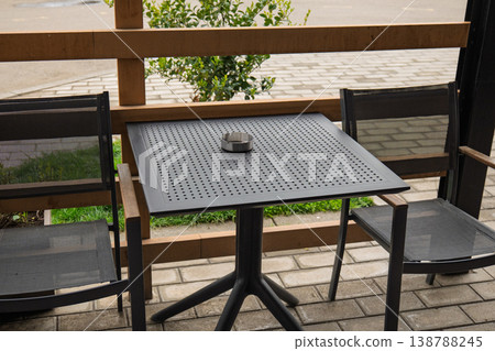 Empty black metal bistro table and mesh chairs on a stone patio with a metal ashtray Empty black metal bistro table and mesh chairs on a stone patio with a metal ashtray 138788245