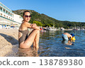 Smiling young adult woman relax bikini sunny beach hold glass wine child play water inflatable arm. Summer vacation lifestyle family carefree leisure mediterranean coast green mountains background 138789380