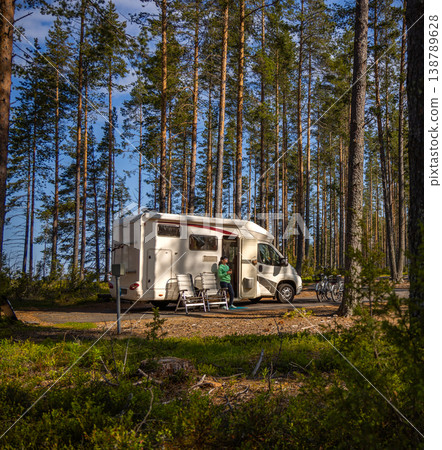 Woman with coffee standing near camper van in pine forest campsite. 138789628