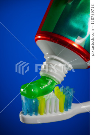 Green gel toothpaste being squeezed from tube onto colorful toothbrush bristles, dental hygiene and oral care concept on bright blue background. Green gel toothpaste being squeezed from tube onto colorful toothbrush bristles, dental hygiene and oral care concept on bright blue background. 138789718
