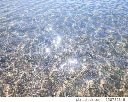 Clean transparent water on shallow sandy seashore with shells with shells on sunny summer day. Sun glare on surface of water. Small waves on sea coast. Nature backdrop. Natural background 138789846