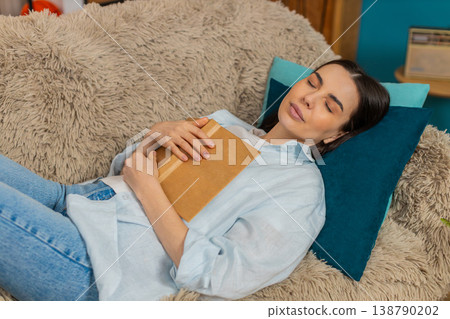 Young woman falling asleep lying on sofa peacefully holding open book tired after reading relaxing Young woman falling asleep lying on sofa peacefully holding open book tired after reading relaxing 138790202