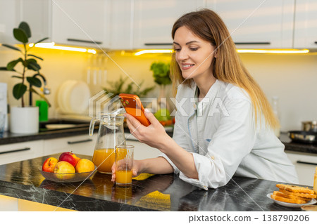Middle-aged woman enjoying smartphone chat while drinking orange juice in kitchen for healthy diet 138790206