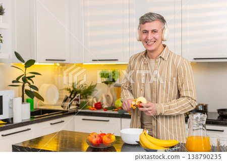 Middle-aged man peeling apple in kitchen with headphones enjoying music during healthy meal routine Middle-aged man peeling apple in kitchen with headphones enjoying music during healthy meal routine 138790235