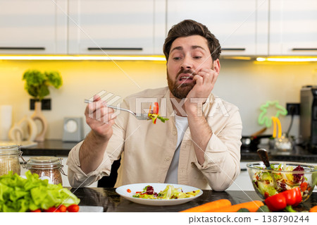 Middle-aged man unhappy eating fresh vegetable salad in home kitchen showing diet willpower struggle 138790244