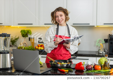 Young woman checking diet recipe on laptop while cooking healthy lunch and writing notes in kitchen 138790267