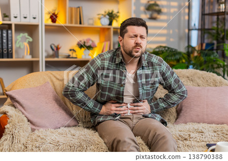 Young man on home sofa suffering sudden stomach pain and trying to ease sharp abdominal discomfort Young man on home sofa suffering sudden stomach pain and trying to ease sharp abdominal discomfort 138790388