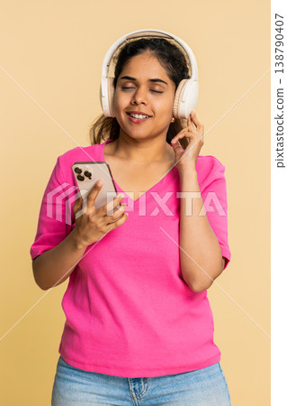 Indian woman relaxing wearing headphones, listening to music on smartphone, gently dancing along Indian woman relaxing wearing headphones, listening to music on smartphone, gently dancing along 138790407