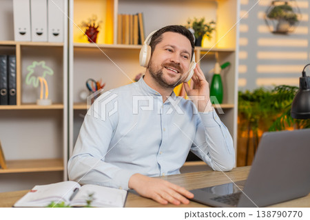 Young man on home table in wireless headphones smiling with music and relaxing after work on laptop 138790770
