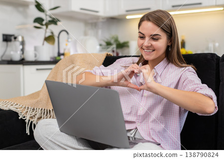 Young woman blogger waves to laptop webcam says hello makes heart sign listens asks chat questions Young woman blogger waves to laptop webcam says hello makes heart sign listens asks chat questions 138790824