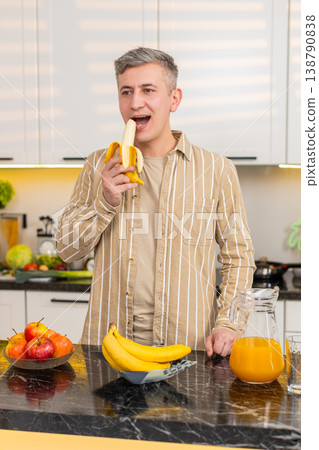 Middle-aged man eating banana in kitchen enjoying healthy eating for weight loss nutrition at home 138790838
