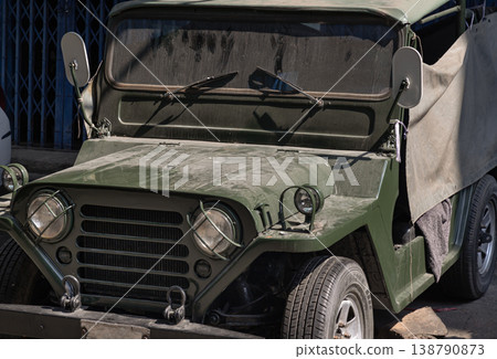 An olive-drab vintage Jeep sits in quiet repose, its caged headlights and weathered canvas top evoking a sense of rugged history against a minimalist urban backdrop. An olive-drab vintage Jeep sits in quiet repose, its caged headlights and weathered canvas top evoking a sense of rugged history against a minimalist urban backdrop. 138790873