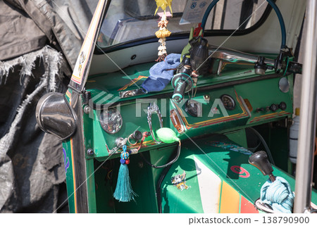 The vibrant green dashboard of this traditional Thai tuk-tuk is a cluttered mix of function and charm, adorned with flower garlands and a swinging turquoise tassel. Sunlight glints off the chrome 138790900