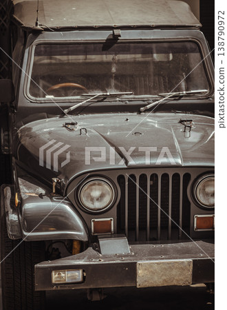 A vintage, dark-colored Jeep sits parked, its classic round headlights and iconic vertical-slotted grille taking center stage.The weathered exterior and aged license plate give the rugged vehicle a A vintage, dark-colored Jeep sits parked, its classic round headlights and iconic vertical-slotted grille taking center stage.The weathered exterior and aged license plate give the rugged vehicle a 138790972