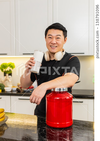Asian man enjoying post workout whey protein drink from shaker cup in cozy home kitchen refuel boost 138791096