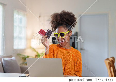 Joyful young African American woman wearing sunglasses holds credit cards while smiling at laptop in cozy home setting. Joyful young African American woman wearing sunglasses holds credit cards while smiling at laptop in cozy home setting. 138791355