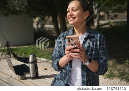 Smiling woman with smartphone working outdoors with laptop, remote work lifestyle Smiling woman with smartphone working outdoors with laptop, remote work lifestyle 138791810