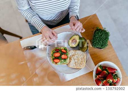 Top view of unrecognizable middle aged woman hands making avocado toast with strawberries and microgreens on bread in home kitchen healthy breakfast preparation natural light lifestyle concept 138792002