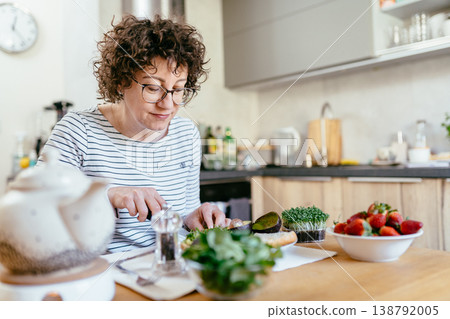 Middle aged woman with curly hair making avocado sandwich in kitchen fresh ingredients strawberries greens morning routine healthy lifestyle natural light 138792005