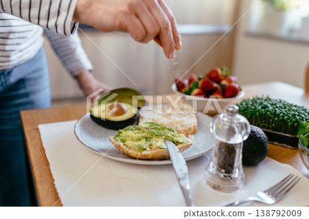 Middle aged woman hand adding salt to avocado toast with microgreens strawberries and tea on table home kitchen healthy breakfast diet control routine Middle aged woman hand adding salt to avocado toast with microgreens strawberries and tea on table home kitchen healthy breakfast diet control routine 138792009