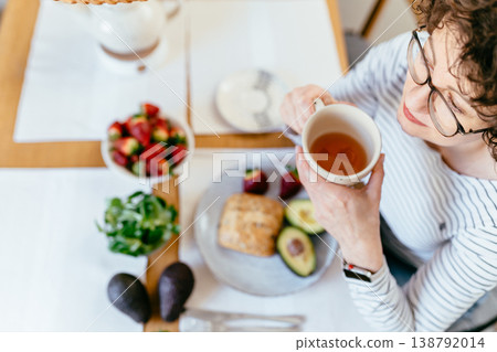 Focused middle aged woman with curly hair and glasses holding cup of tea over healthy breakfast with avocado bread strawberries greens and microgreens top view home kitchen wellness routine Focused middle aged woman with curly hair and glasses holding cup of tea over healthy breakfast with avocado bread strawberries greens and microgreens top view home kitchen wellness routine 138792014