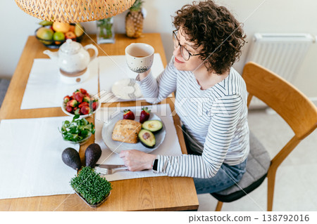 Relaxed middle aged woman with curly hair and glasses sitting at table enjoying tea and healthy breakfast with avocado bread strawberries greens and microgreens cozy home morning routine.. Relaxed middle aged woman with curly hair and glasses sitting at table enjoying tea and healthy breakfast with avocado bread strawberries greens and microgreens cozy home morning routine.. 138792016